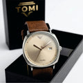 Watch with brown leather strap and gold face on a black box with 'TOMI' branding.
